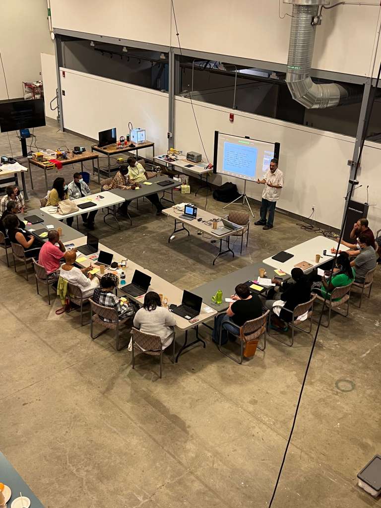 Overhead view of a large, open room where a group of people are seated around a U-shaped arrangement of tables, participating in a workshop or training session. A presenter stands at the front near a projection screen, and laptops, notebooks, drinks, and cables are spread across the tables. The industrial-style space features high ceilings, exposed ductwork, and concrete floors.