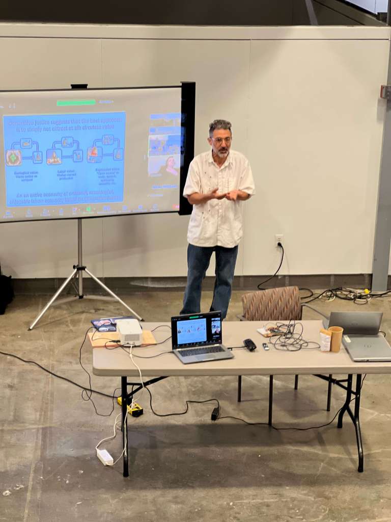 A man stands in front of a projection screen giving a presentation in an open room with concrete floors. He is wearing a white shirt and jeans, gesturing with his hands. In front of him is a table with laptops, cables, a coffee cup, and presentation equipment. The projected slide appears to feature diagrams and text with a blue background.