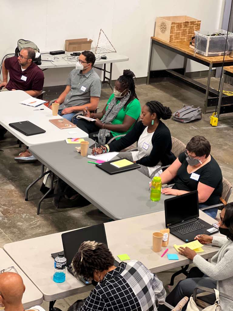 A diverse group of people seated around two rectangular tables arranged in a U-shape, engaged in a collaborative meeting or workshop. Most participants have laptops, notebooks, or sticky notes in front of them. Several individuals are wearing face masks, and coffee cups and water bottles are visible on the tables. The room has a concrete floor and industrial-style furnishings.