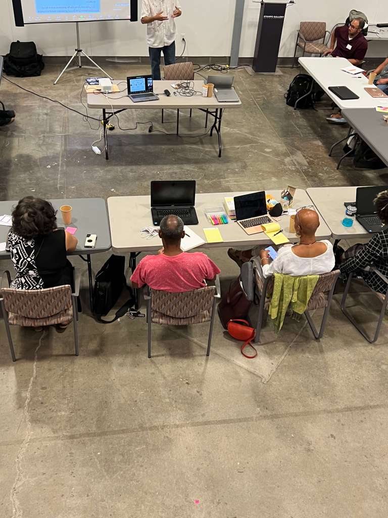 Overhead view of a group of people seated at tables arranged in a U-shape, participating in a presentation or workshop. Laptops, notebooks, drinks, and colorful sticky notes are on the tables. A presenter stands at the front near a projection screen, while participants listen and take notes in an open room with concrete floors.