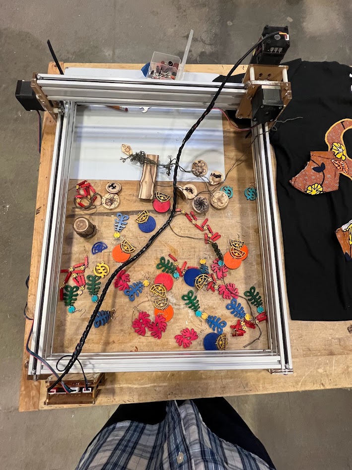 Overhead view of a laser cutter or CNC machine on a wooden table, filled with colorful cut-out shapes made from wood or acrylic, including flowers, leaves, and abstract designs. The machine is connected with wires and components, and to the side is a black T-shirt with applique pieces. A person in a plaid shirt is partially visible at the bottom of the image, suggesting an active maker or craft workshop setting.