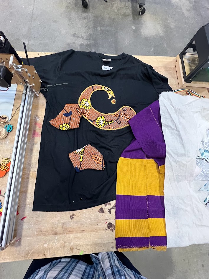 A black T-shirt with colorful, patterned applique cutouts arranged on it, laid out on a wooden worktable alongside a purple and yellow knitted garment and part of a white fabric item. The appliques include floral and abstract shapes in shades of orange, yellow, and brown. The setting appears to be a craft or maker space, with tools and equipment visible in the background.