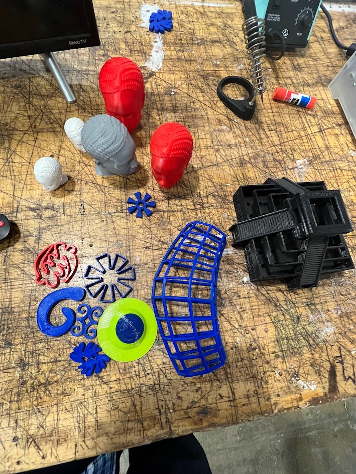 A cluttered wooden workbench covered with various colorful 3D printed objects, including stylized heads in red and gray, abstract geometric shapes in blue, green, and red, and black stackable components. A computer monitor, glue stick, and other tools are partially visible, indicating an active maker or fabrication workspace.