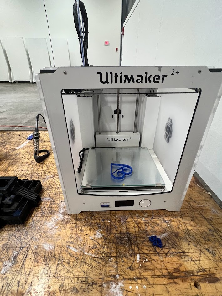 Alt text: An Ultimaker 2+ 3D printer sits on a well-used wooden workbench, actively printing a blue object shaped like a stylized symbol or logo. The surrounding area shows signs of creative use, with filament scraps and tool cases nearby. The scene appears to be in a makerspace or workshop environment.