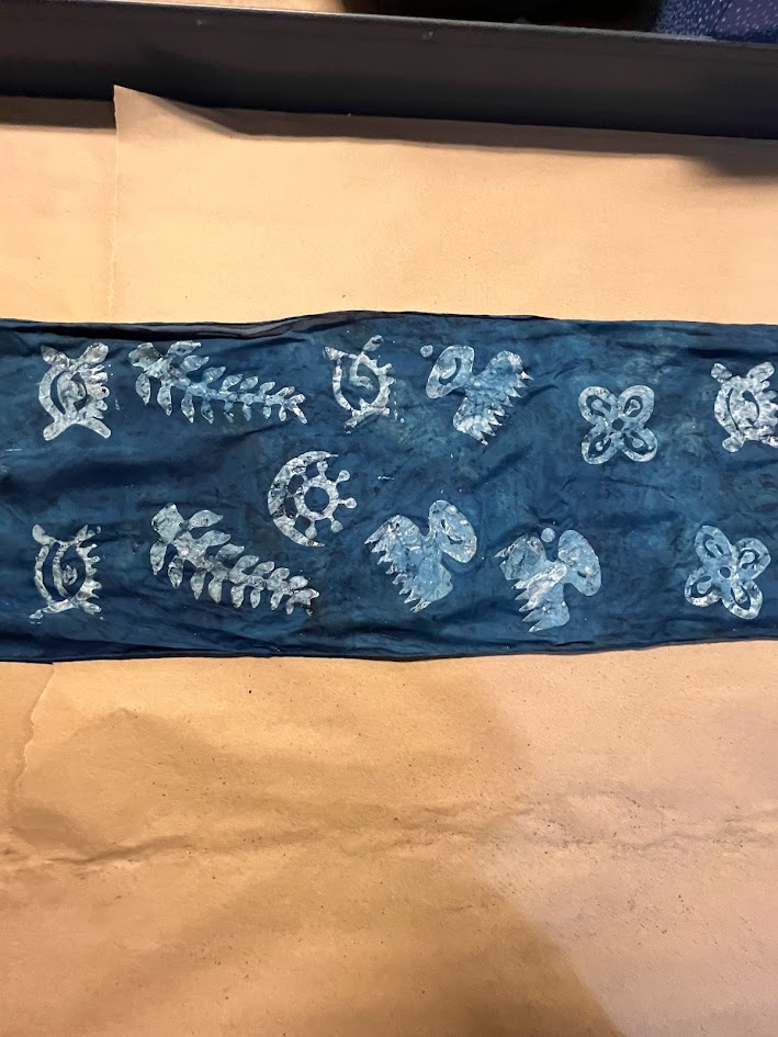 A strip of dark blue fabric dyed with white or light blue traditional-style motifs, including ferns, birds, triskelions, and eye-like symbols. The fabric is laid flat on a piece of brown paper, likely as part of a textile art or dyeing workshop. The designs appear to be created using a resist or stencil technique.