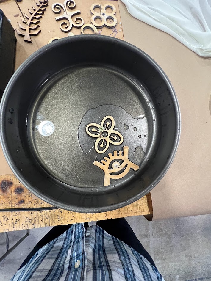 A round metal pot with a small amount of water at the bottom contains two decorative wooden pieces—one shaped like a triskelion and the other resembling an eye with lashes. The pot is placed on a wooden surface, and more laser-cut wooden symbols are visible nearby. The lower part of a person wearing a plaid shirt is seen standing in front of the setup.