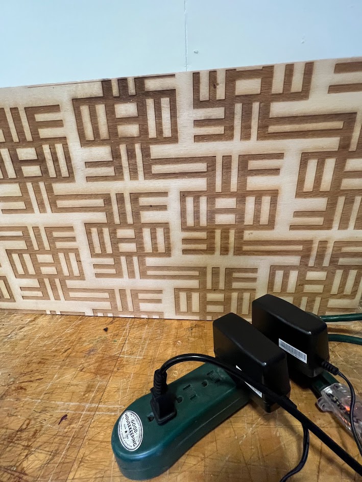 A close-up of a wooden surface featuring a laser-etched geometric pattern made up of interlocking rectangular shapes. In front of the panel is a green power strip with electronic plugs and black adapters connected to wires, resting on a scratched wooden table.