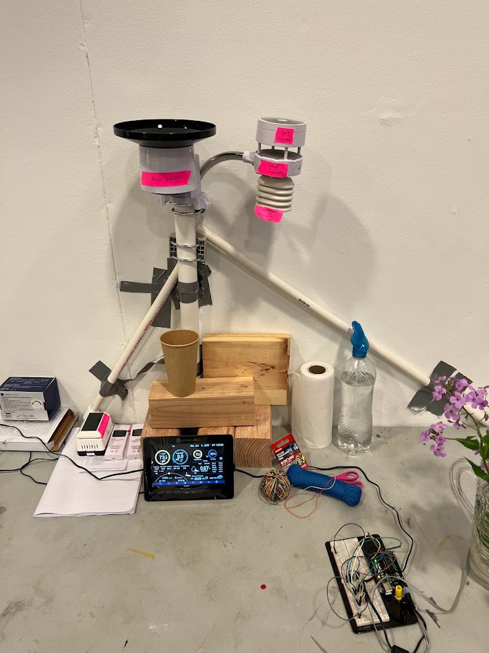 A DIY weather station setup displayed against a wall, featuring labeled sensors for rain, light, and temperature mounted on PVC pipes. Below the sensors, various components are arranged on the floor and a wooden base, including a tablet displaying sensor data, an electronic breadboard circuit, a multimeter, a spray bottle, and a paper towel roll. A vase with flowers and boxes of electronic components are also part of the scene, suggesting an experimental or educational environment.