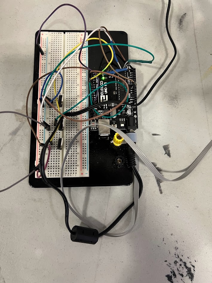 A close-up of an electronic breadboard project with multiple colored jumper wires connected to an Arduino microcontroller. The setup is placed on a black platform on a table, with various cables and connectors arranged around it. Some faint smudges are visible on the tabletop surface.