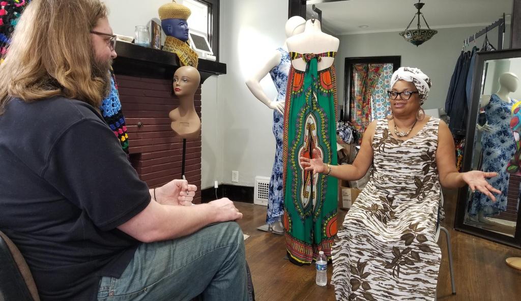A researcher with long hair and glasses interviews a Black woman artisan in a colorful textile studio. The woman, wearing a patterned dress and headwrap, gestures expressively while mannequins dressed in vibrant African fabrics and a mannequin head on a stick surround them. The setting is warm and creative, highlighting handmade fashion and cultural craftsmanship.